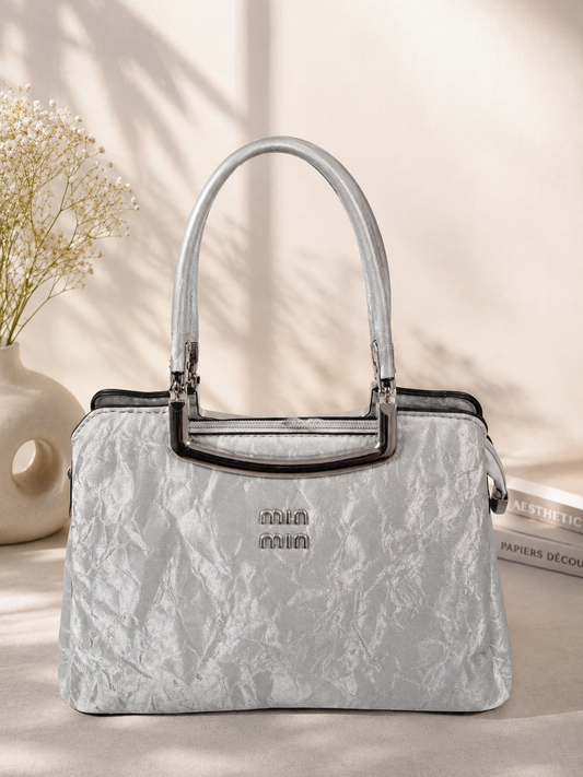 A silver textured handbag with a structured dome shape, double top handles, and a small silver logo on the front, styled on a beige surface with soft shadows, a vase of baby's breath, and books in the background.